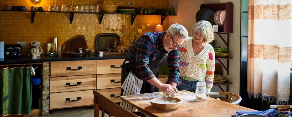 A retired couple roll out bread dough together on their dining table.