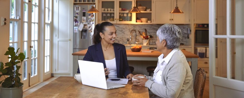 A woman discusses finances with an advisor at her kitchen table, with documents and a laptop in front of them.