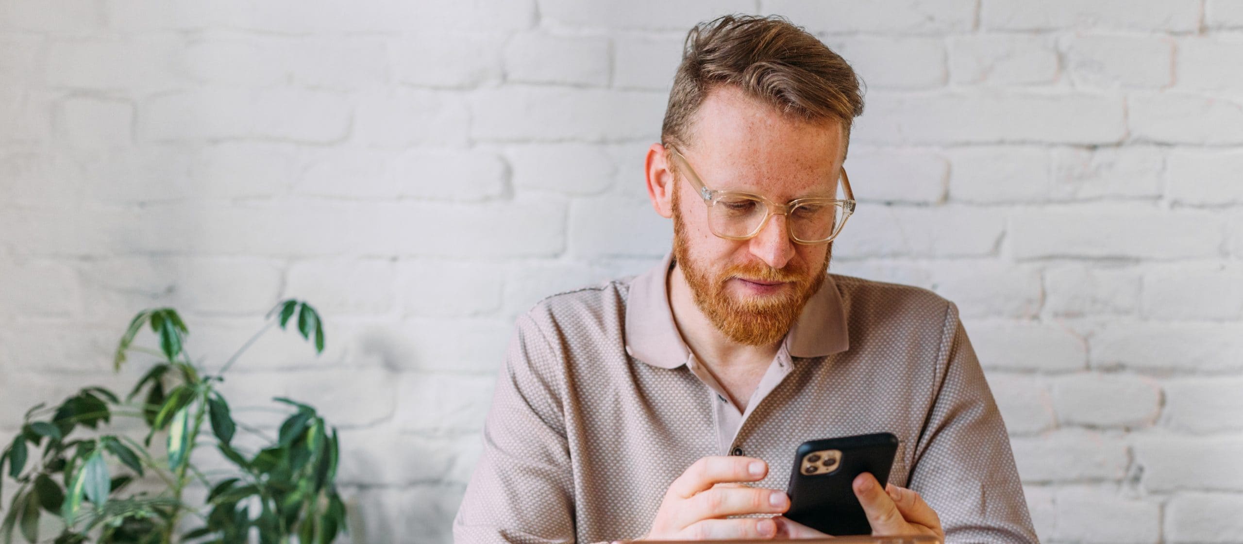A man sits and looks at his
phone, with a plant and white
brick wall in the background.