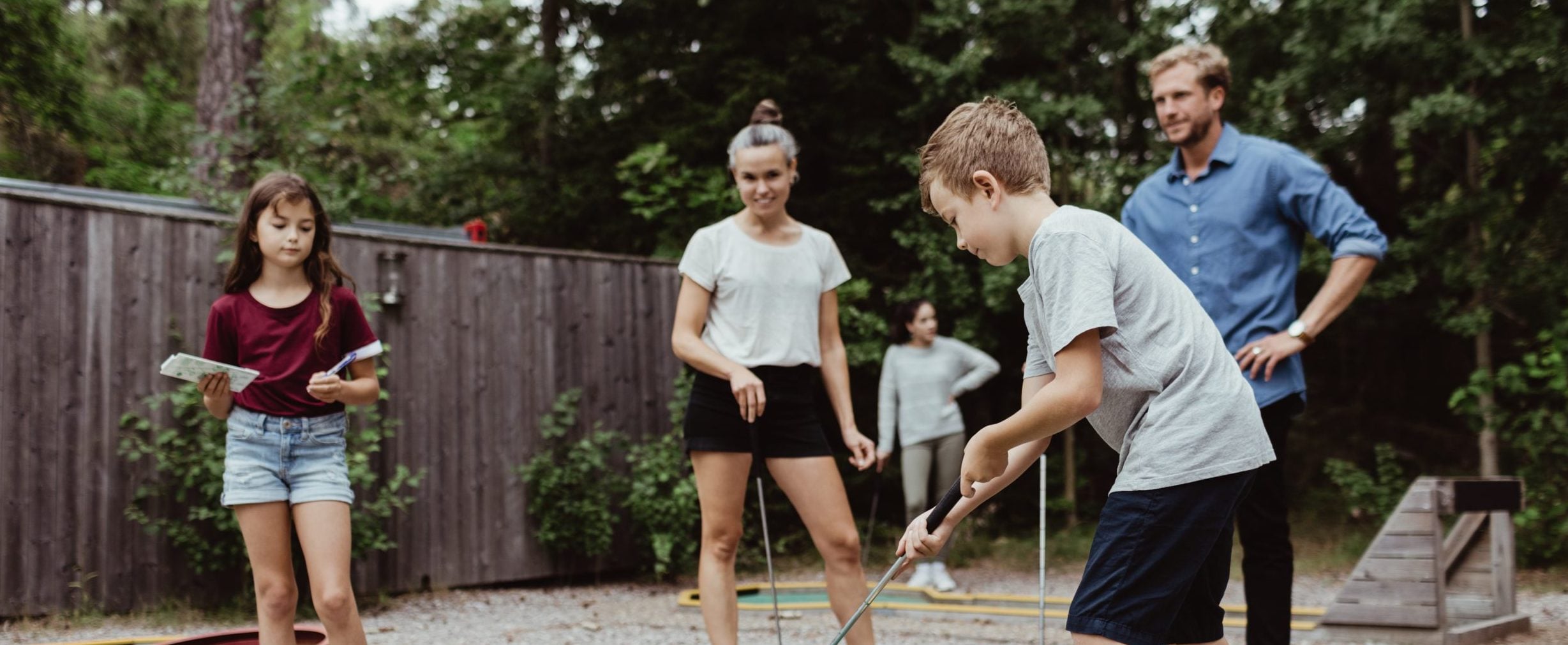 Parents watching their son play miniature golf, while their daughter is holding a scorecard and a pen.