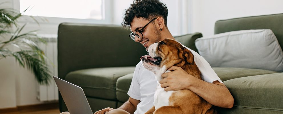 A young man sits on the floor with his laptop, hugging his dog and smiling.