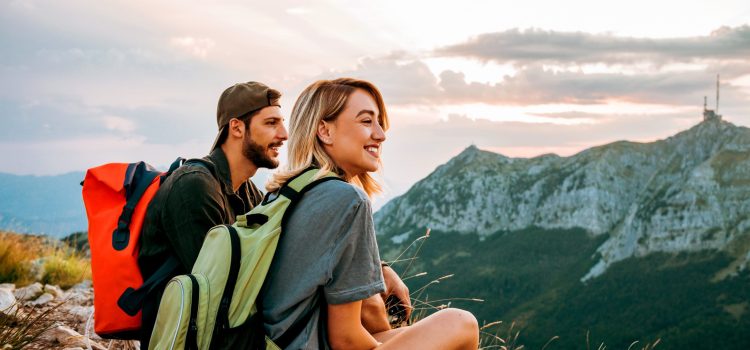 A man and woman are sitting, wearing backpacks and look out over a mountainous landscape