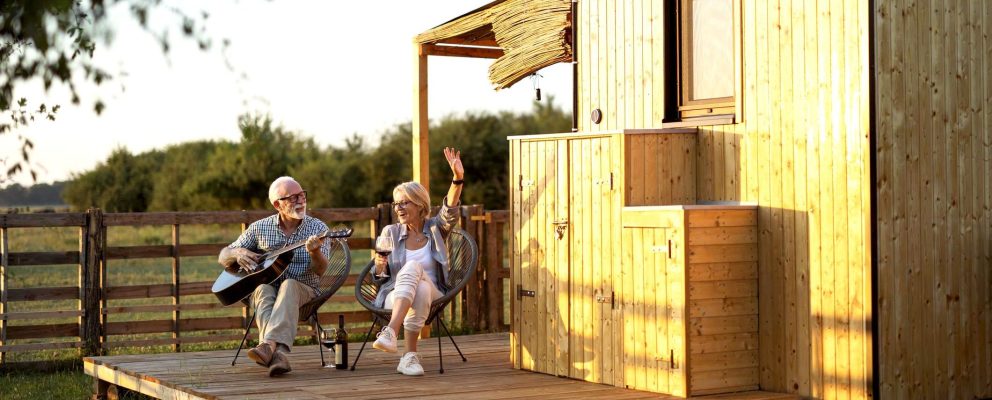 A retired couple sits on chairs outside. The man plays a guitar while the woman sings and drinks wine.